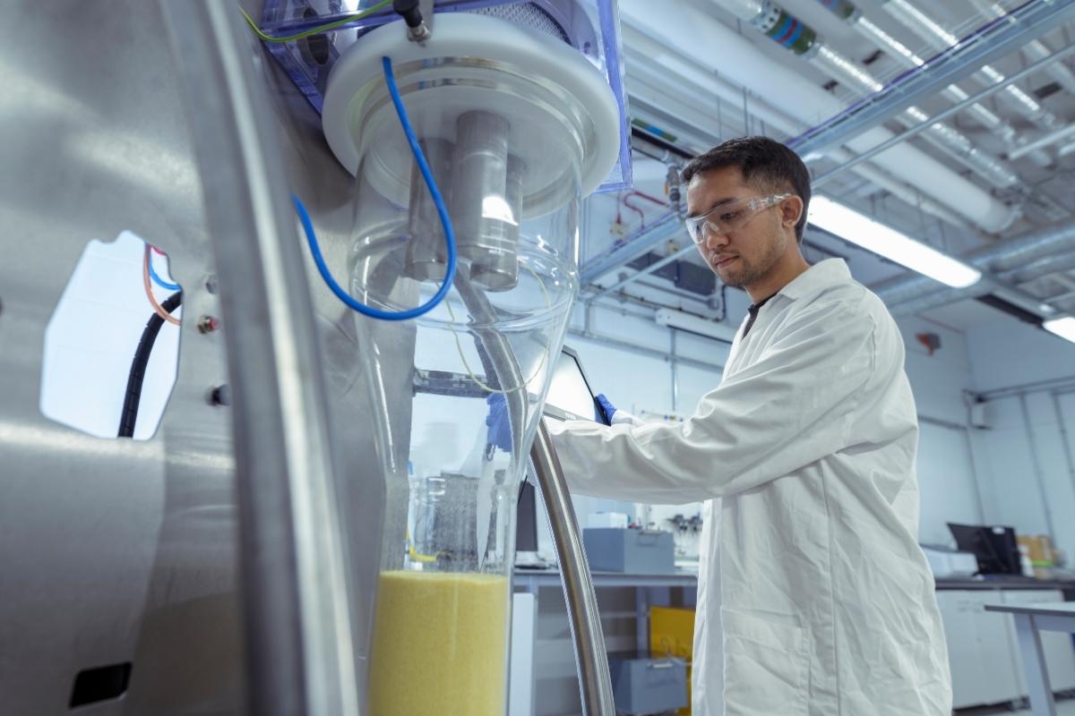 A student is wearing a white lab coat and protective eyewear, working with specialist equipment in a lab.