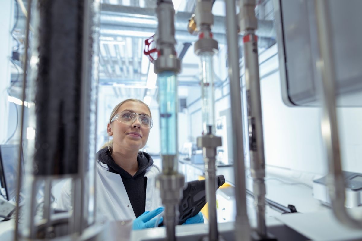 A student is wearing a white lab coat and protective eyewear, working with specialist equipment in a lab.