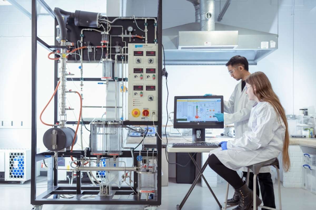 Two students are wearing white lab coats and working with a large piece of specialist equipment looking at a computer monitor.