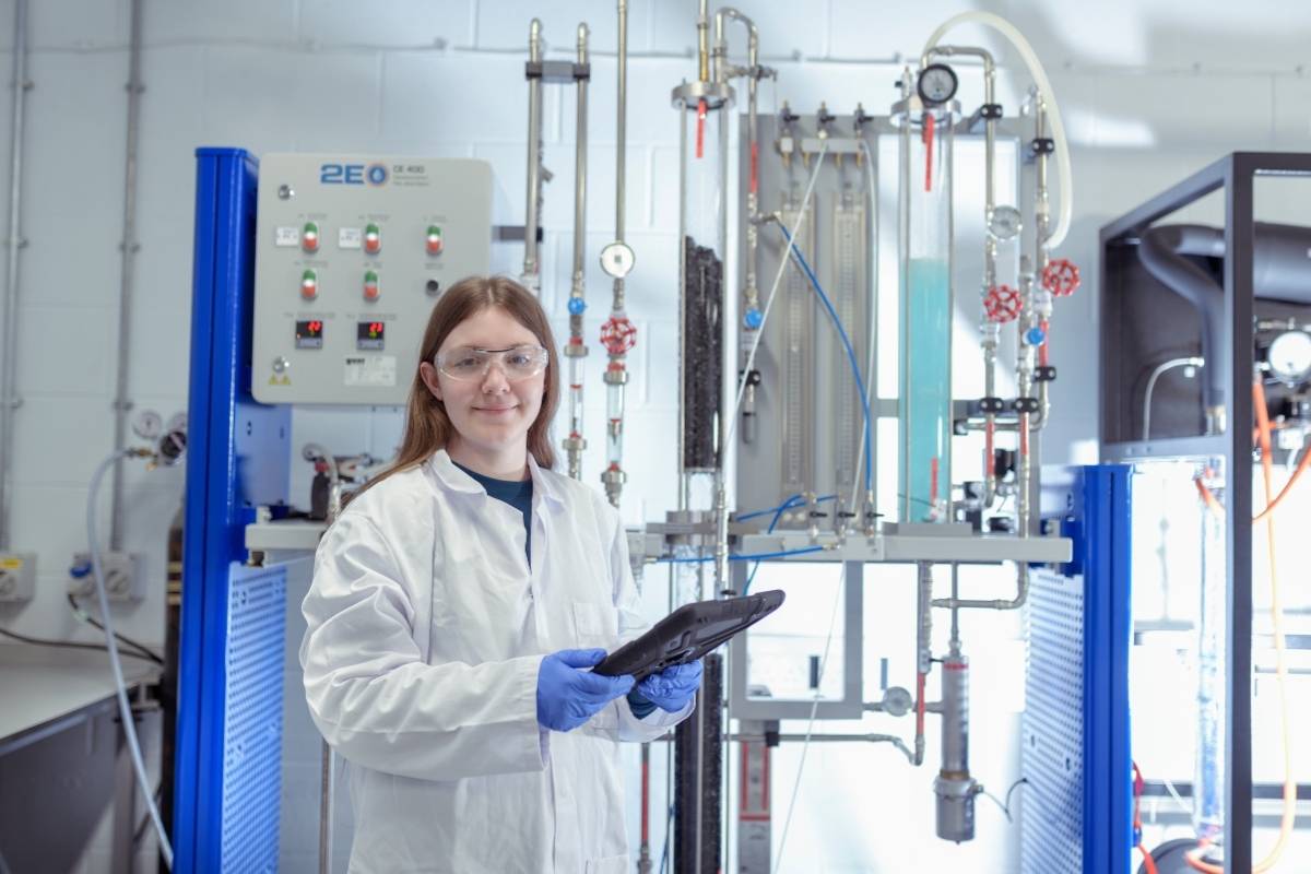 A student is holding a tablet and wearing a white lab coat, blue rubber gloves and protective eyewear with specialist engineering equipment in the background.