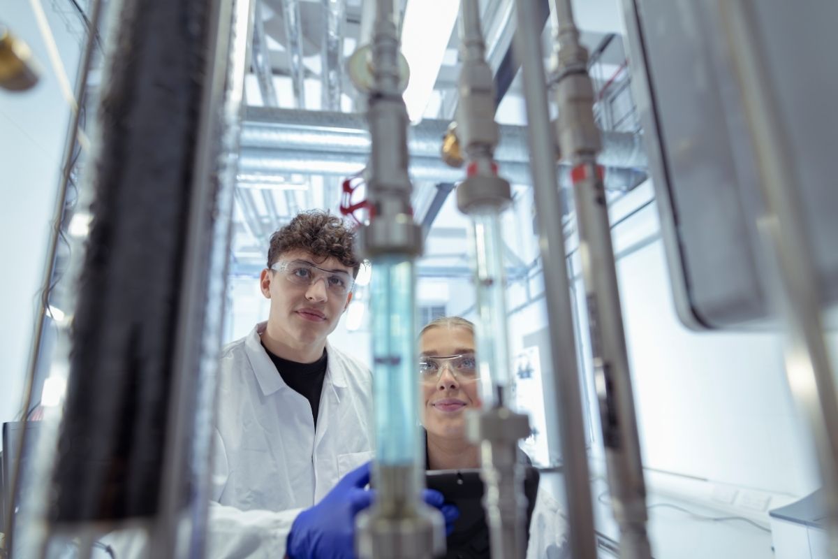 Two students are wearing protective eyewear and white lab coats inspecting equipment that has metal pipes and glass tubes.