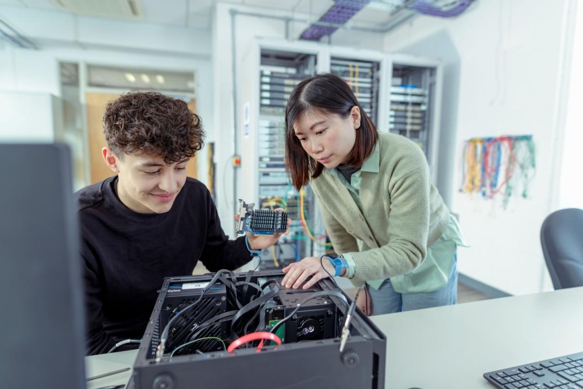 A tutor is assisting a student with a piece of electrical equipment on a table in a brightly lit workshop space.