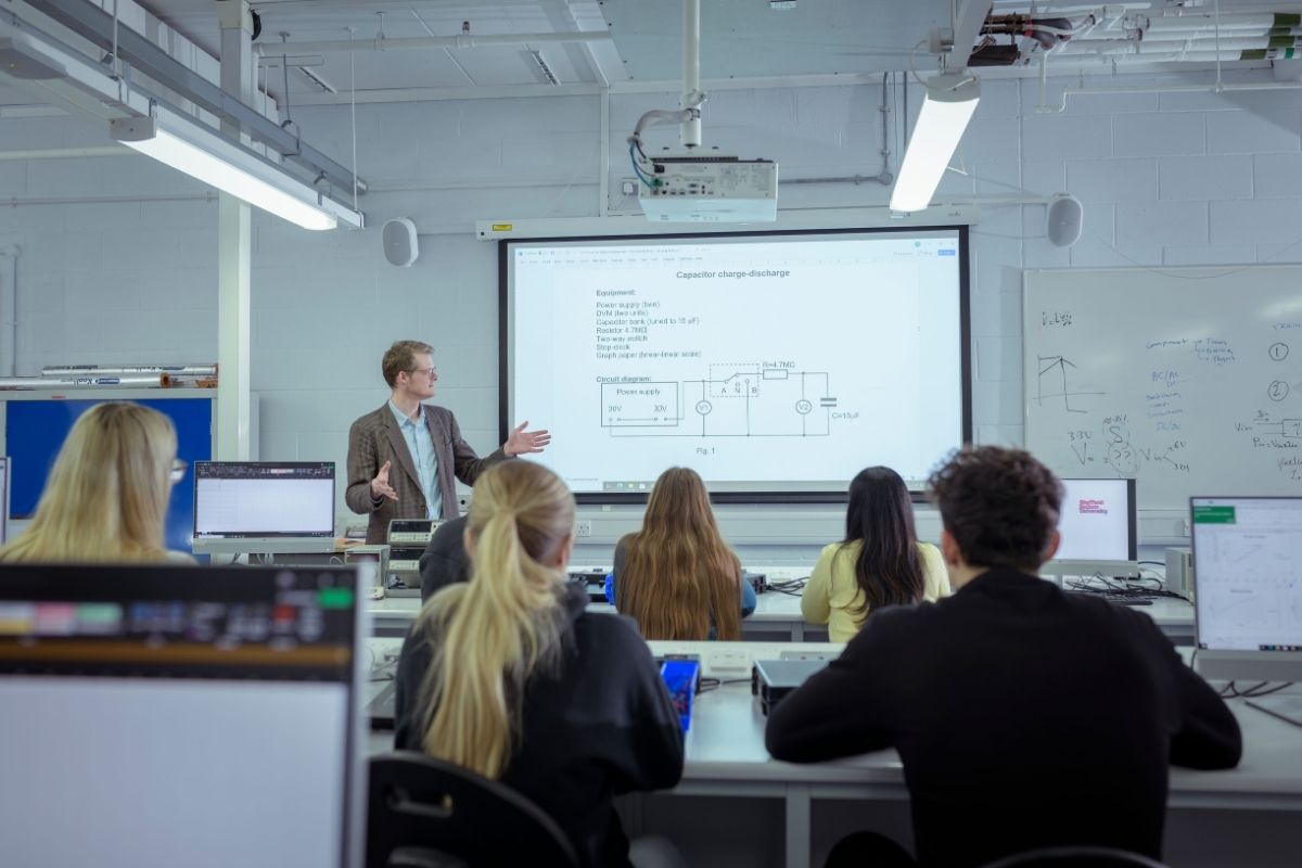 A lecturer is talking to students in a lecture room and is stood next to a digital board, which is displaying text and charts.