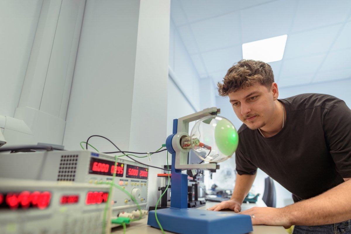 A student is leaning over a table to look at physics equipment in a brightly lit lab.