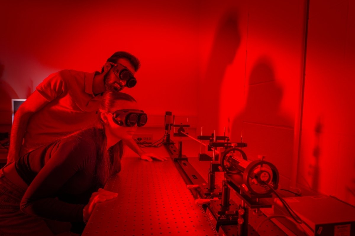 Two students are stood in a dark-room wearing protective eyewear and looking at physics equipment.