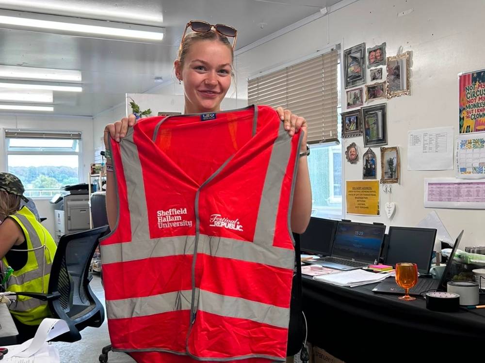 Abbie is stood in a small office holding up a hi-vis vest with Sheffield Hallam and Festival Republic logos. In the background, there are computer monitors, lots of paperwork and posters.