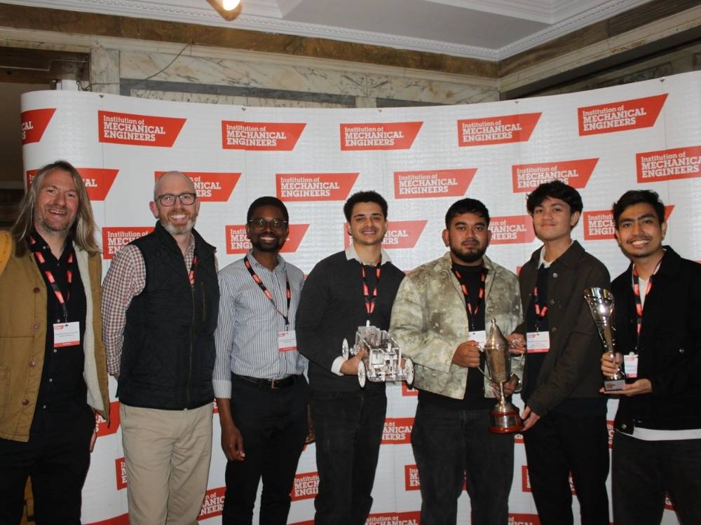 Four engineering students are holding two silver trophies and a small robotic vehicle, stood in front of a promotional board displaying the Institution of Mechanical Engineers orange logo.