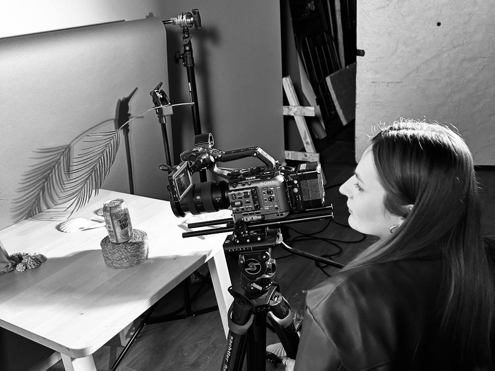 Charlotte Turnock is using a high spec camera to take a photograph of a can of tropical drink next to a backdrop of tropical leaves and shells.