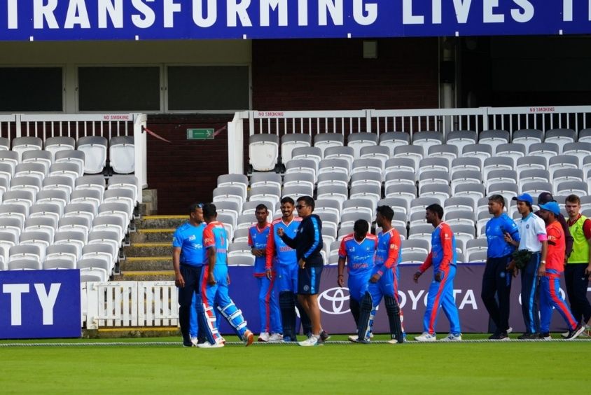 Group of male cricket players on a cricket pitch. Kashish is in the middle in a dark outfit talking to a cricket player
