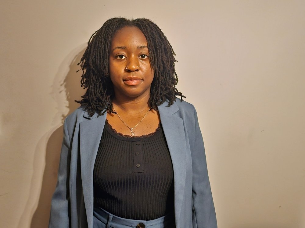 Malikah has a calm expression and is wearing a grey blazer over a black t-shirt.