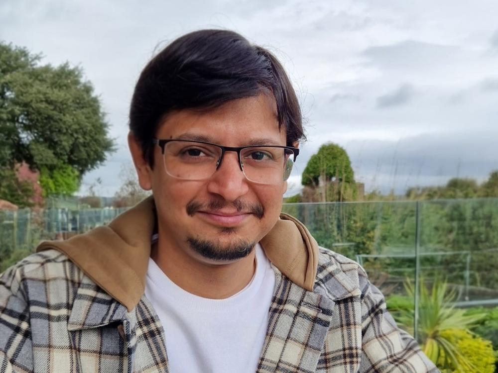 Rohan is smiling and wearing eye glasses, a white t-shirt and checked jacket, standing outside with trees and plants in the background.