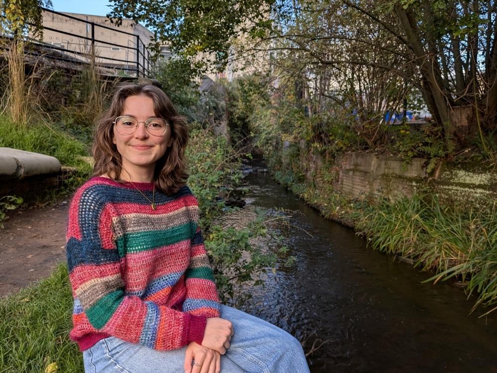 Ruth is wearing a colourful striped jumper and jeans, sat on a grassy bank next to a stream, with trees and buildings in the background.