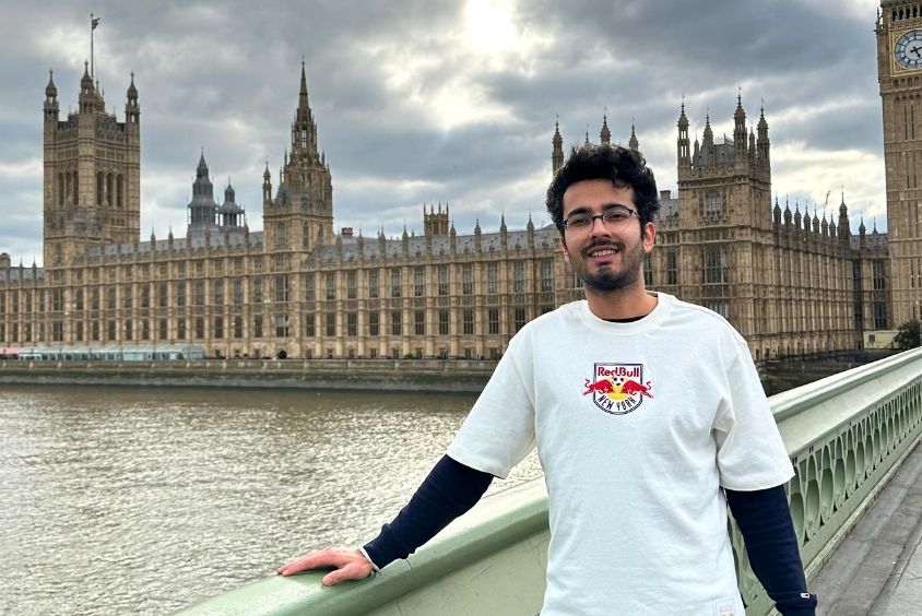 Man wearing glasses and a white t-shirt, standing on a bridge with the view Westminister, London in the background