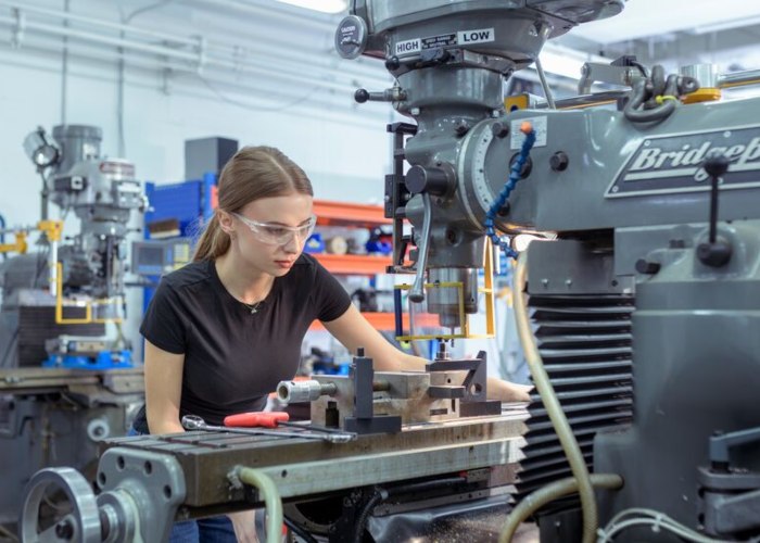 A student girl wearing safety glasses and operating a large drill machine in a Sheffield Hallam workshop.