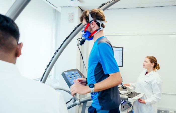 A person running on a treadmill wearing a breathing mask while a technician monitors data on a nearby computer.