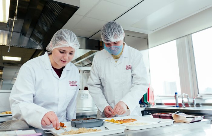 Two people in lab coats and hairnets preparing food samples on a stainless steel counter.