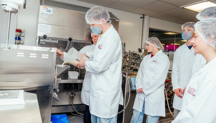A group of people in lab coats and hairnets observing a machine while one person pours liquid into it.