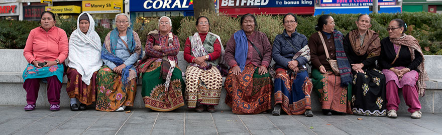 A group of Nepali women seated on a wall on a high street, with shops in the background. The image is a still from the film Alone Together.