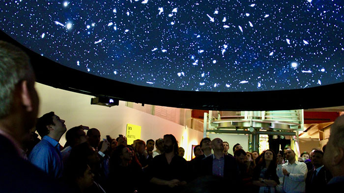A group of people in an art gallery. They are looking upwards at stars projected onto a large black canvas above them. 