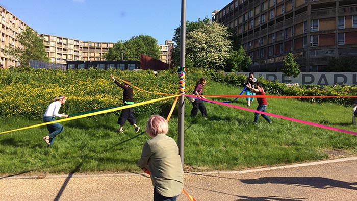 A group of artists winding coloured ribbons round a streetlight in Sheffield's Park Hill estate. There are estate blocks in the background, and in the foreground a group of six people is winding ribbons around a pole at a distance. There is an expanse of grass behind them, and the sun is out.