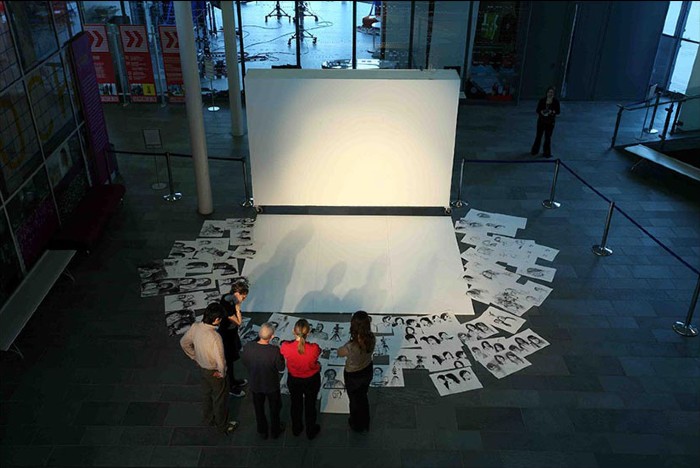An image from the Arrivals and Departures: New Art Perspectives of Hong Kong exhibition. A group of people huddled around a series of images on the floor and a large blank canvas.