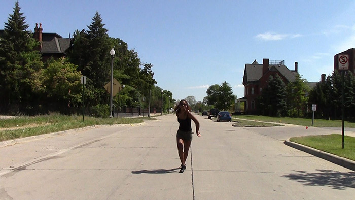 A woman dancing alone in the middle of a suburban street. The street is long, wide and straight, with wide grass verges either side. There is a patch of tall trees in the background on the left, and a large house can be seen behind them.