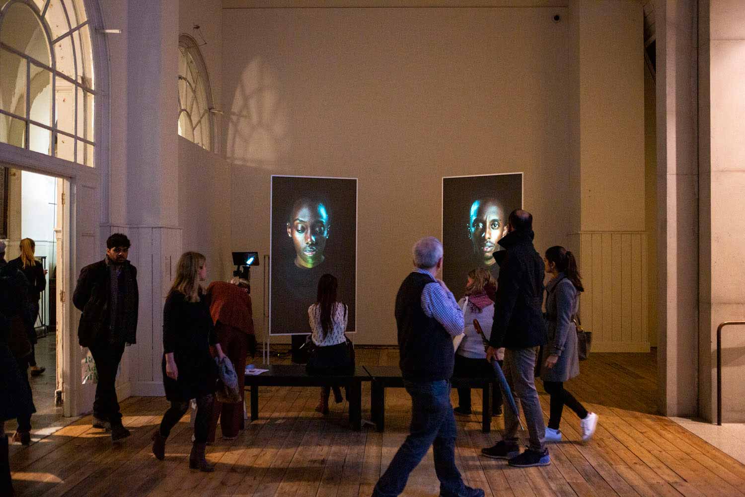 Installation image of Mirror IV by David Cotterrell. Two people are shown on screens in front of a group of people in a gallery.