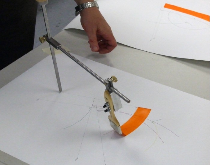 A compass-like contraption with a paint brush on the end, painting an arch over the top of some drawings on paper.