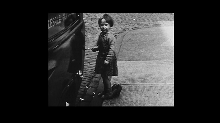 A black and white still from the From Scotland with Love film. A little girl stands next to a car