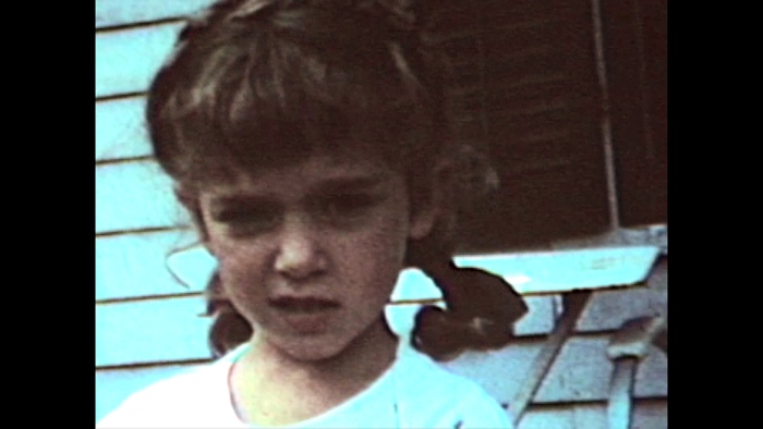 Vintage photo of a young girl. She is half smiling and has pigtails. Close behind her is the wall of a clapboard house.