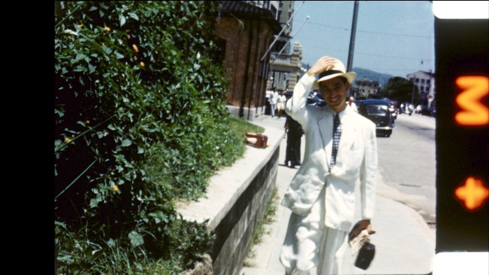 A man in a white suit on a city street. He is about to raise his hat with his right hand. The image looks old, and the border has markings that suggest it was developed from film, not taken digitally,