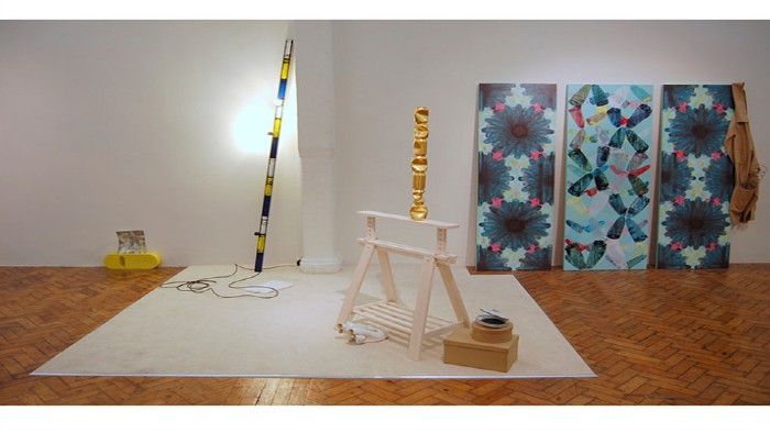 A piece of art from the Pile exhibition. A white sheet covers the wooden floor with a wooden white sculpture and gold ornament on top. Three blue and pink patterned painting in the background. 
