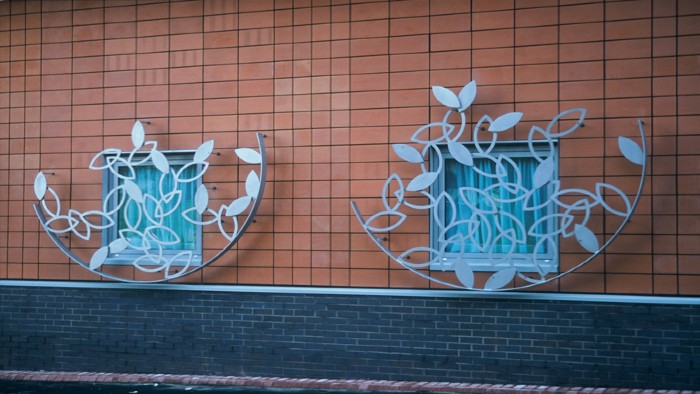 A piece of metalwork on the side of a city centre building. The metalwork is a semicircle shape filled with solid and outlined leaves.