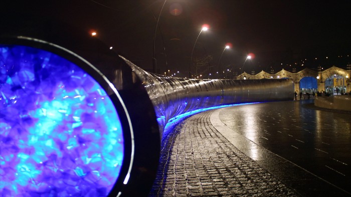 The metalwork water fountain erected outside of the Sheffield Station, the centre of it lit up with a blue mottled light at night. The photograph is taken looking toward Sheffield station