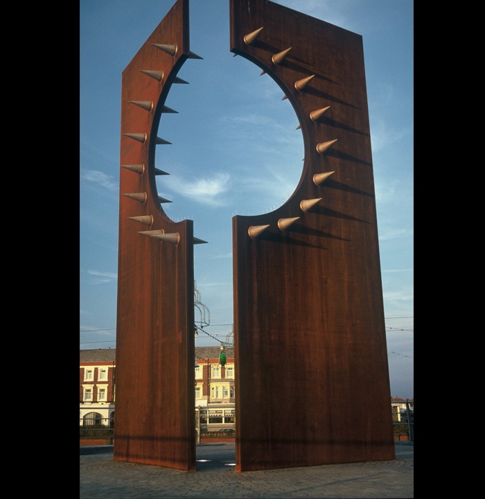 An erected piece of metalwork. Two tall pieces of metal stand perpendicular to each other each with a semicircle cut out surrounds by pointed studs