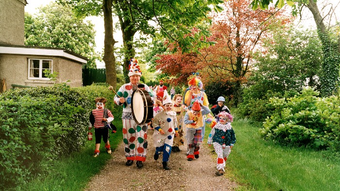 A group of adults and children dressed up as clowns walking down a gravel path with a building on the left and a green forest area on the right