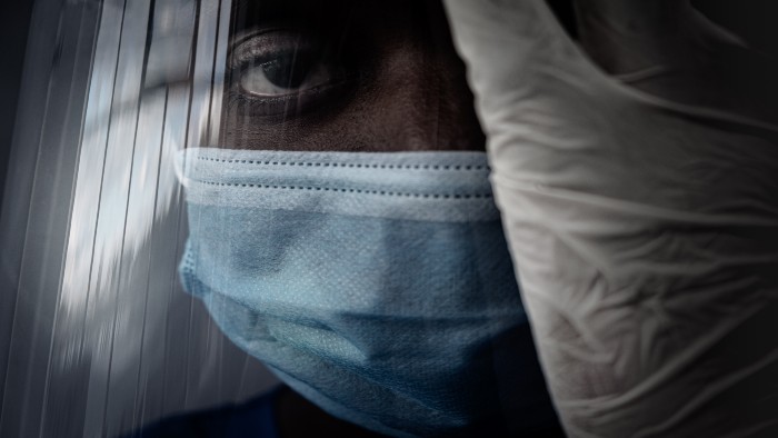Face of a black healthcare worker, wearing a blue mask and a clear face shield with a latex gloved hand covering the right side of their face