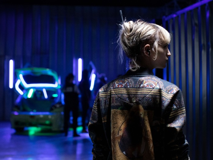 A woman in the right foreground looking to the right. In the background with people next to a car with a the boot open surrounded by neon lights