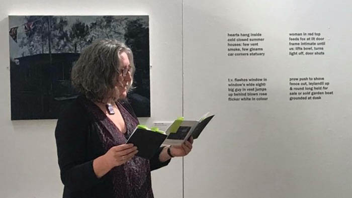 Professor Harriet Tarlo giving a poetry reading. She is standing in front of a white wall, on which are displayed a black and white photo and four stanzas of poetry. She is holding a small concertina book, and looking down at it as she reads.