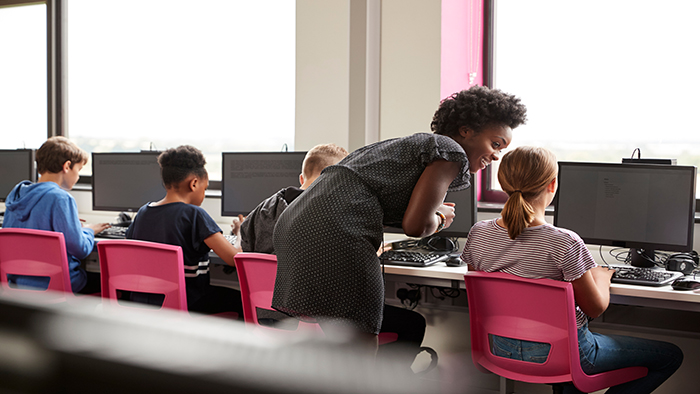 Female Teacher Supervising Line Of High School Students Working at Screens In Computer Class
