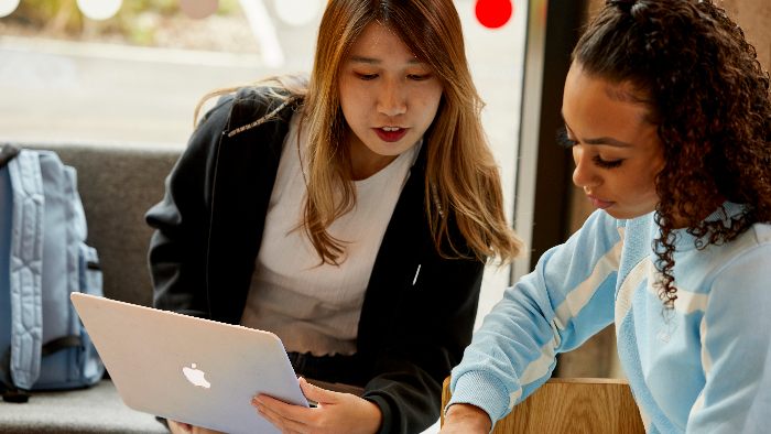 Two students sat next to each other on a communal sofa. One is helping the other with some work from a laptop.