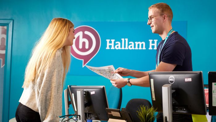 A person leans over a helpdesk speaking to another person holding a map. Behind them is a blue wall that says 'Hallam Help' and on the desk are PC monitors.