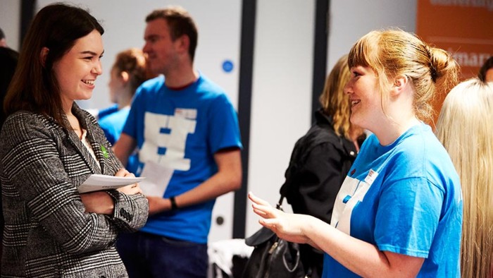 Two people talking at a Sheffield Hallam open day event, one holding a notepad, the other in a blue open day ambassador t-shirt.