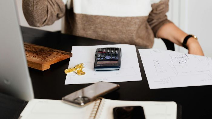 A person is sitting at a desk with a calculator, house keys, papers, a phone, and a notebook in front of them.