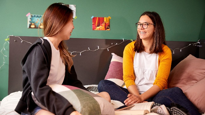 Two students sat cross legged on a bed smiling and talking surrounded by several large patterned cushions.