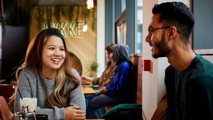Two students sat at a table in a coffee shop talking and smiling. A milkshake and tea infuser are on the table.