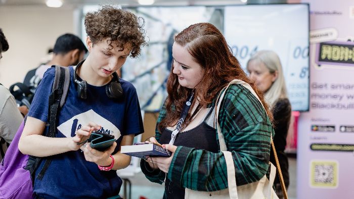 Two students are looking at a phone together at an event. One is holding a phone and the other is holding a notebook. There are other people and display stands in the background.