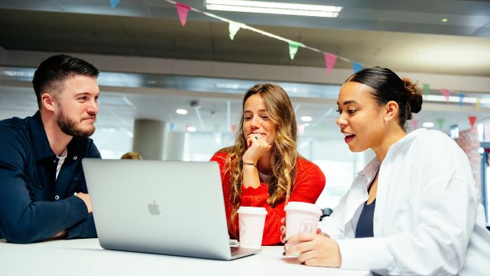 Three people at a table around a shared laptop, holding cups and talking, under colourful hanging flags in a bright indoor space.