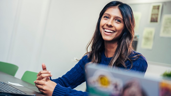 A smiling person in a blue sweater sits at a desk with laptops in a bright office or classroom setting.