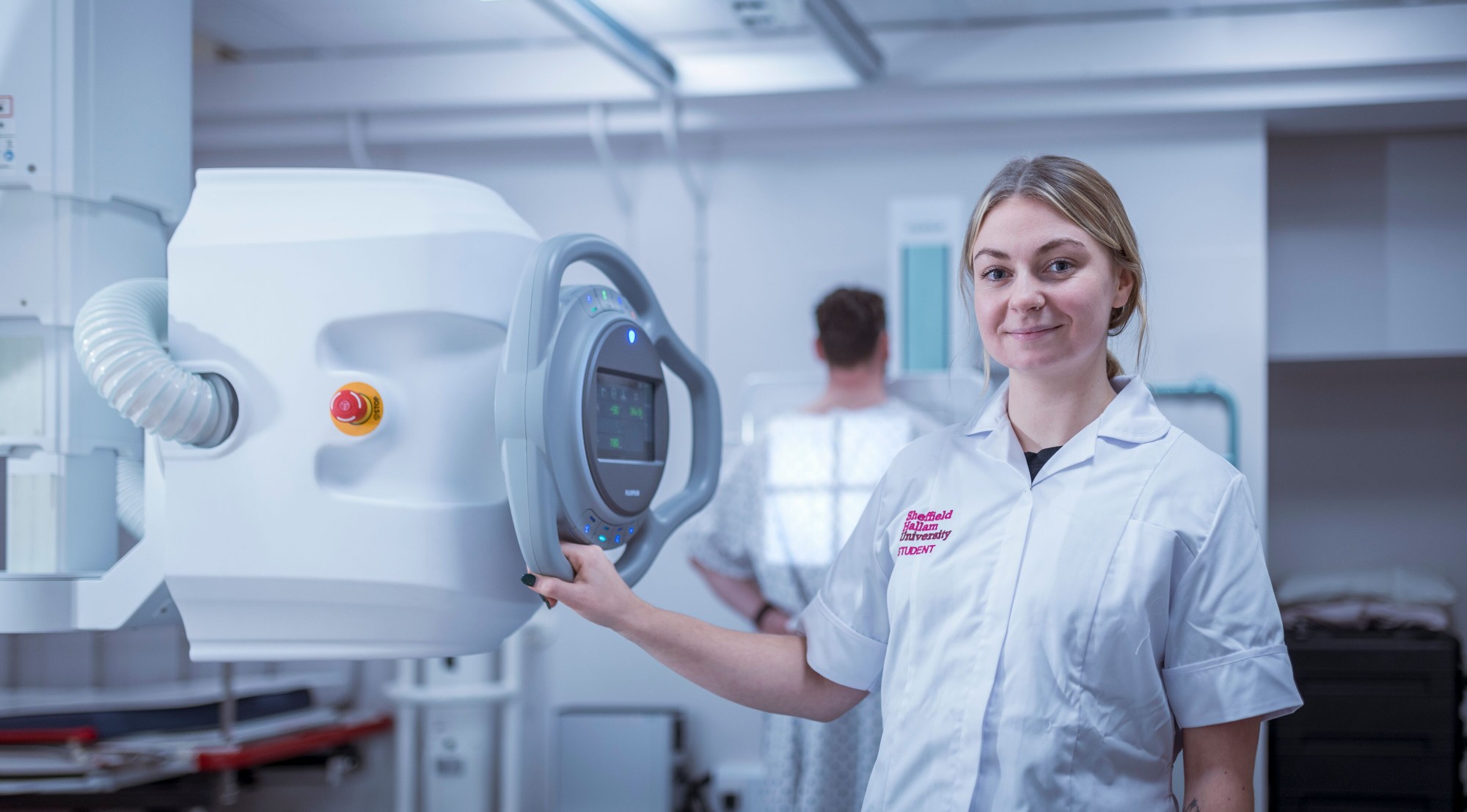 A Sheffield Hallam student stood smiling for a photo. She is wearing a medical uniform and holding onto medical apparatus.